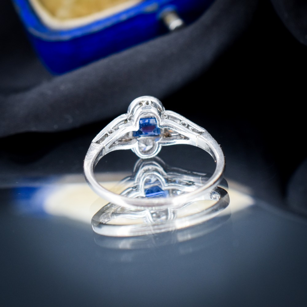 An Art Deco platinum ring showcasing a central rectangular step-cut blue sapphire framed by small round diamonds set in a geometric, elongated mounting. View VI