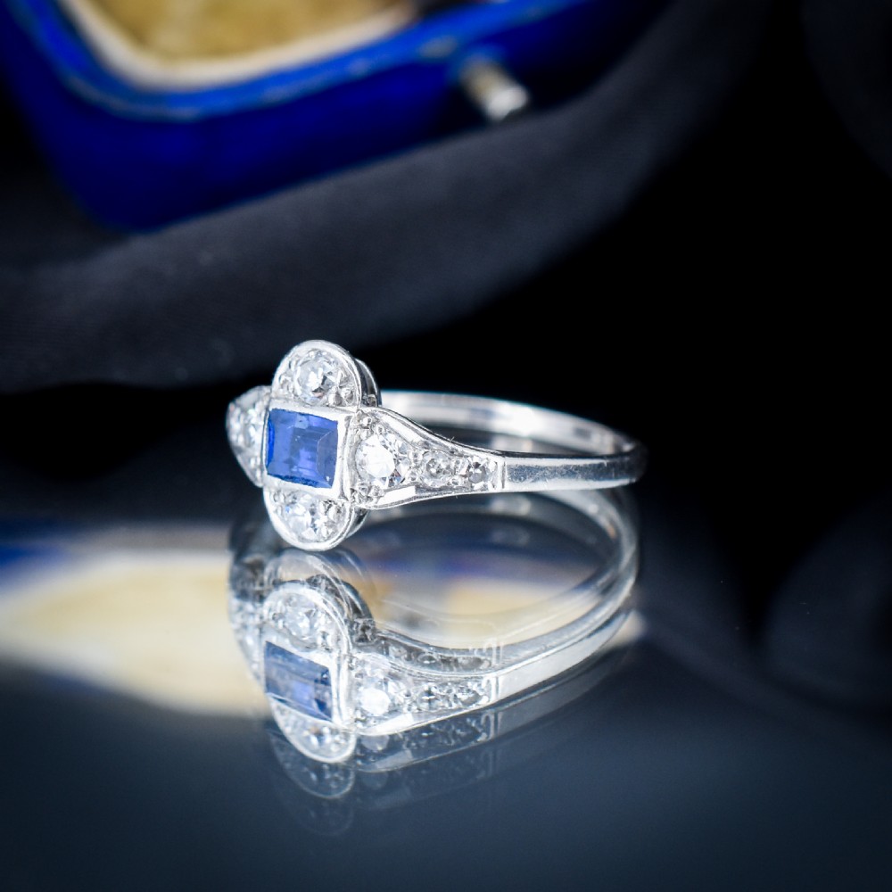 An Art Deco platinum ring showcasing a central rectangular step-cut blue sapphire framed by small round diamonds set in a geometric, elongated mounting. View VII