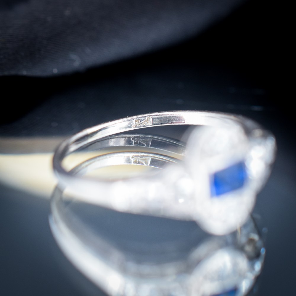 An Art Deco platinum ring showcasing a central rectangular step-cut blue sapphire framed by small round diamonds set in a geometric, elongated mounting. View VIII