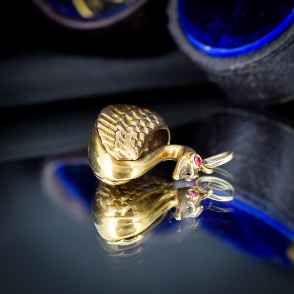 A small 9ct yellow gold swan charm featuring textured feather details, a round ruby set as the eye, and visible hallmarks stamped on the side. View VI