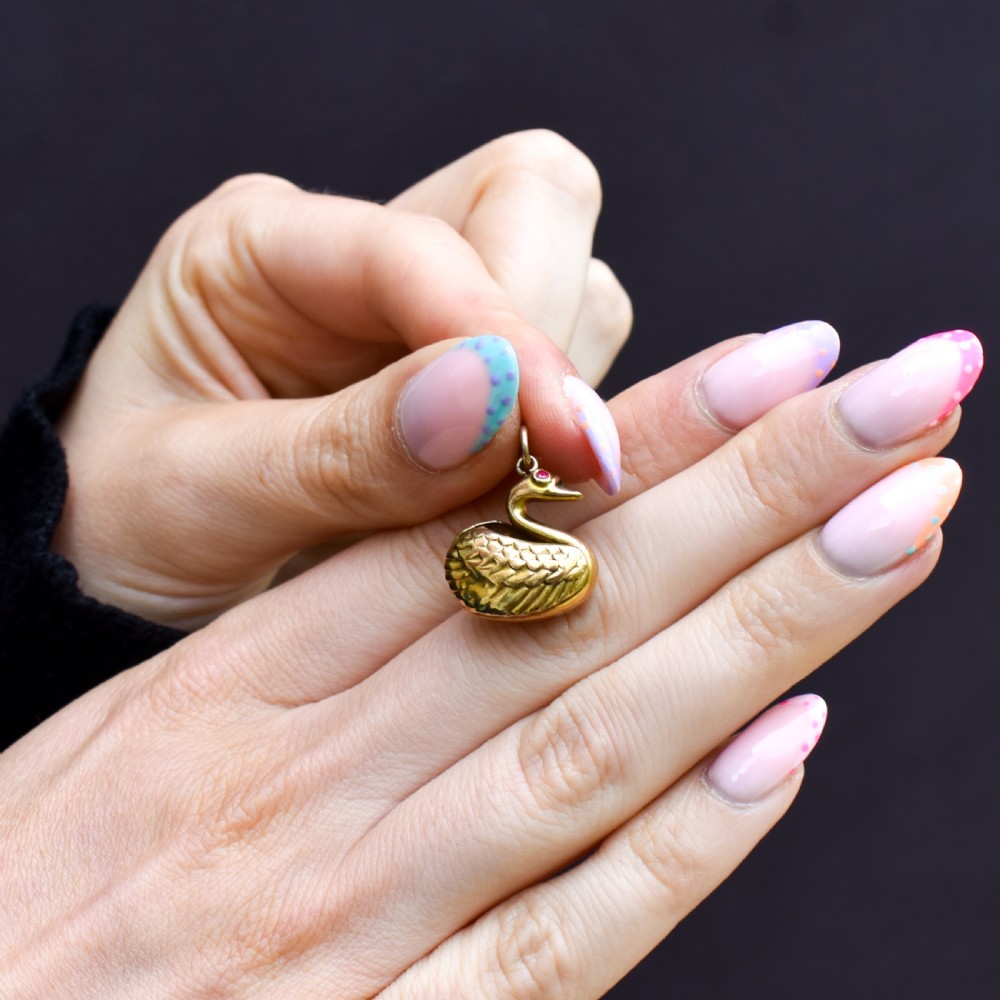 A small 9ct yellow gold swan charm featuring textured feather details, a round ruby set as the eye, and visible hallmarks stamped on the side. View VII