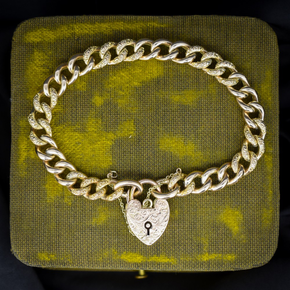 A 9ct yellow gold curb link chain bracelet featuring alternating plain and hand-engraved links, fitted with an engraved heart-shaped padlock clasp and safety chain.