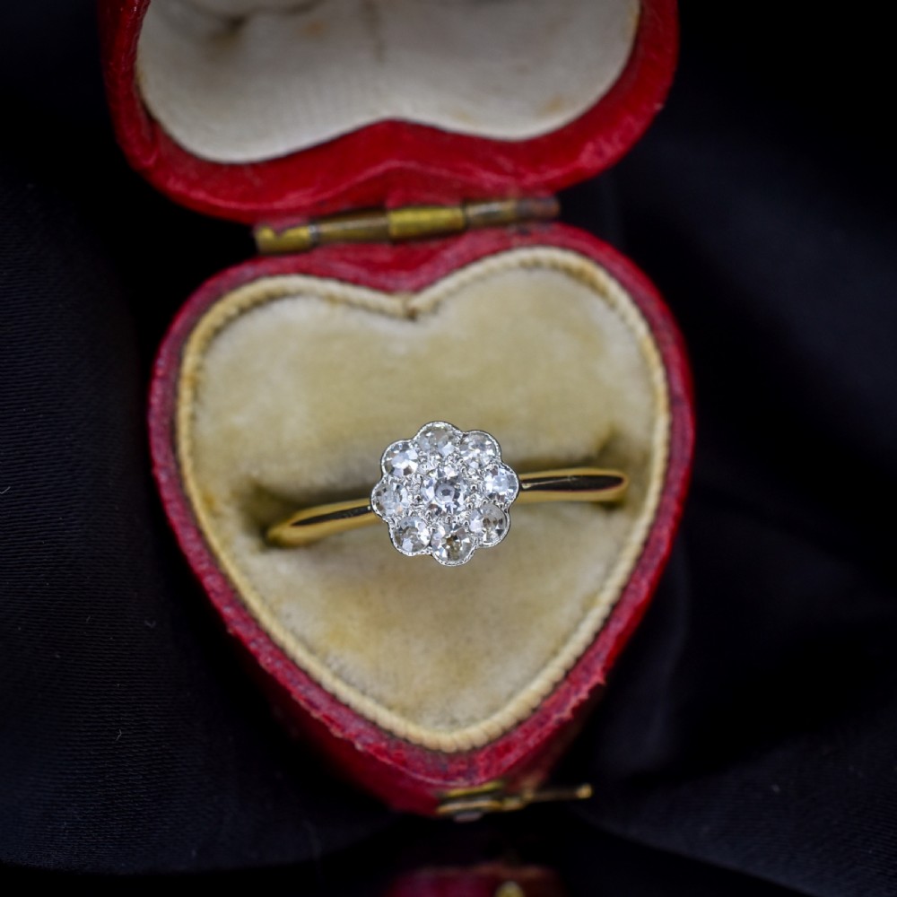 An antique diamond daisy cluster ring set in platinum with an 18ct yellow gold band resting inside an open red heart-shaped velvet box.