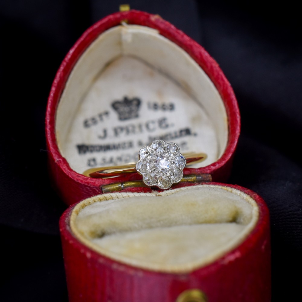 An antique diamond daisy cluster ring set in platinum with an 18ct yellow gold band resting inside an open red heart-shaped velvet box. View V