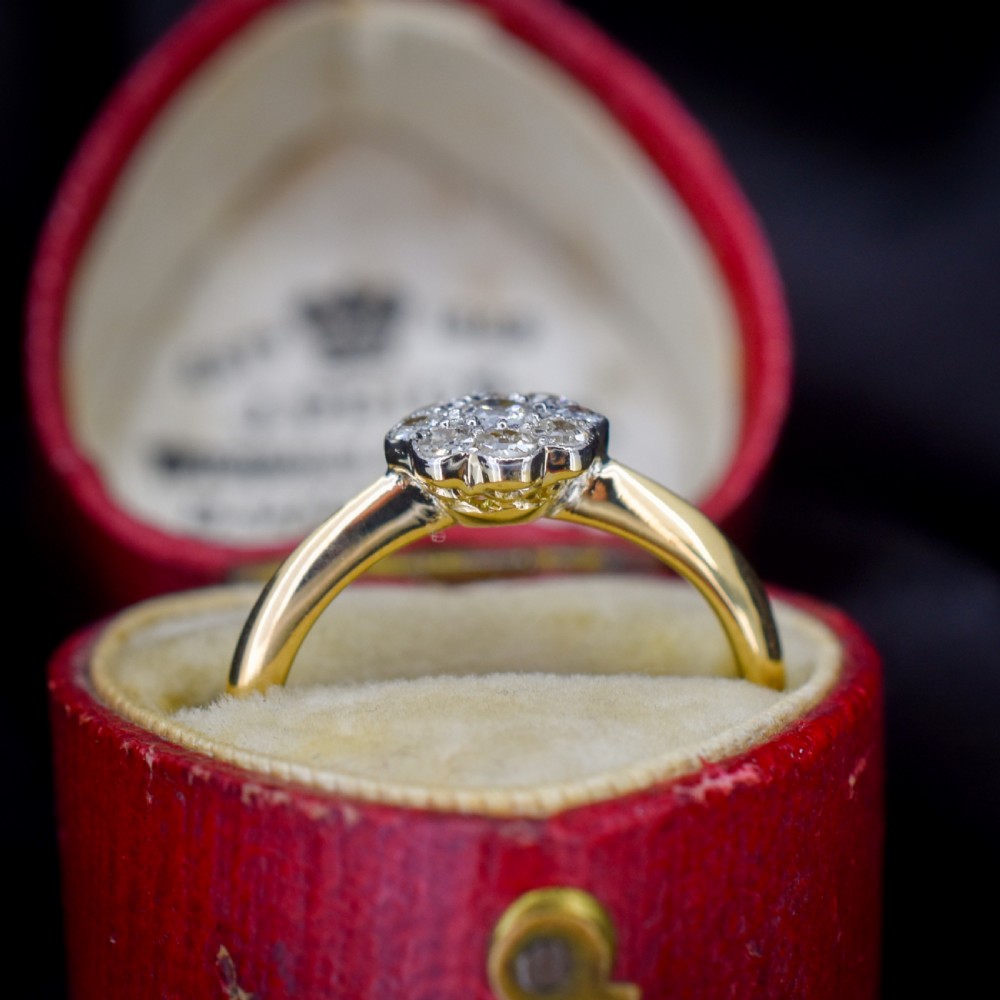 An antique diamond daisy cluster ring set in platinum with an 18ct yellow gold band resting inside an open red heart-shaped velvet box. View VI