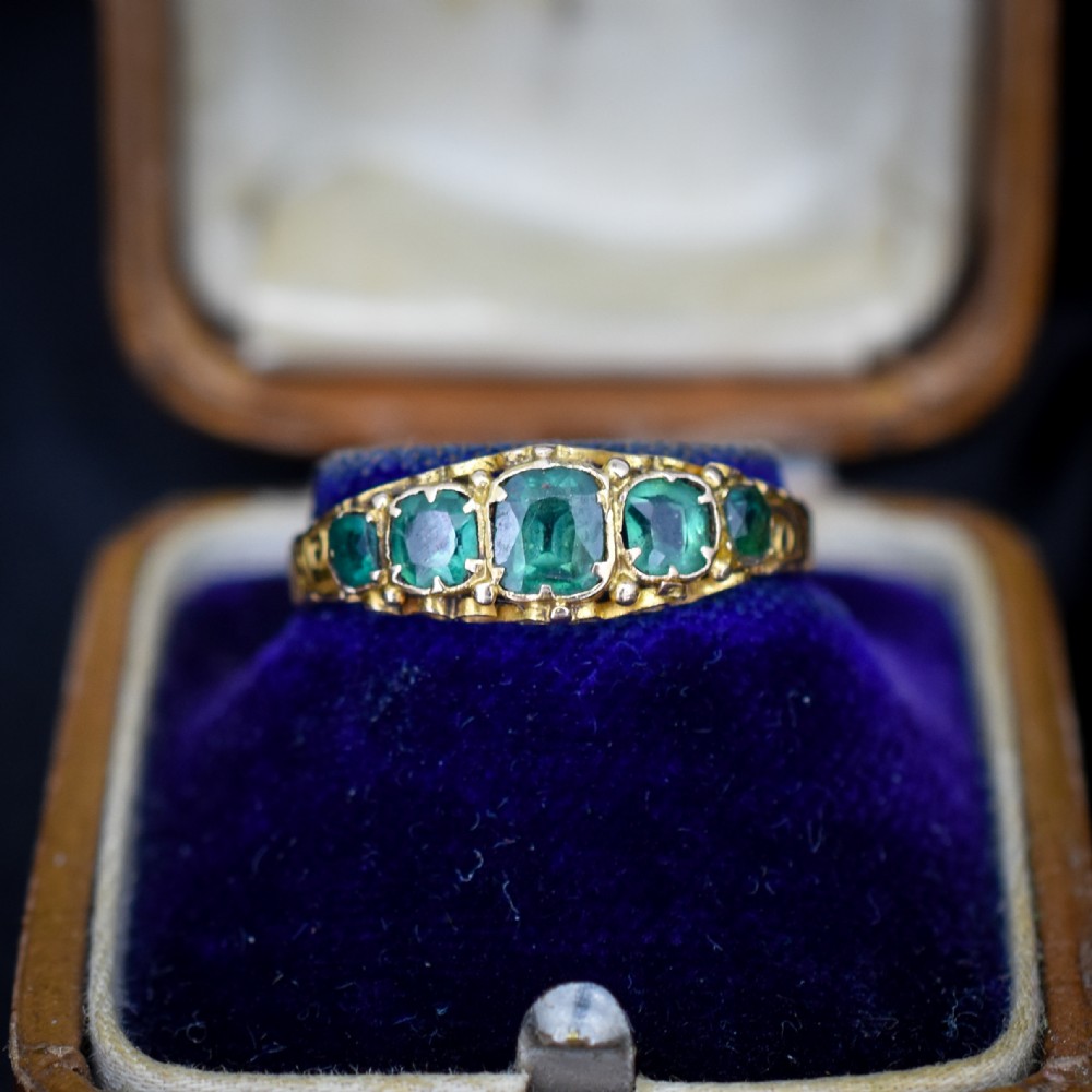 An antique Victorian 15ct yellow gold ring featuring five graduated cushion-cut emerald green paste stones set with carved scroll shoulders.