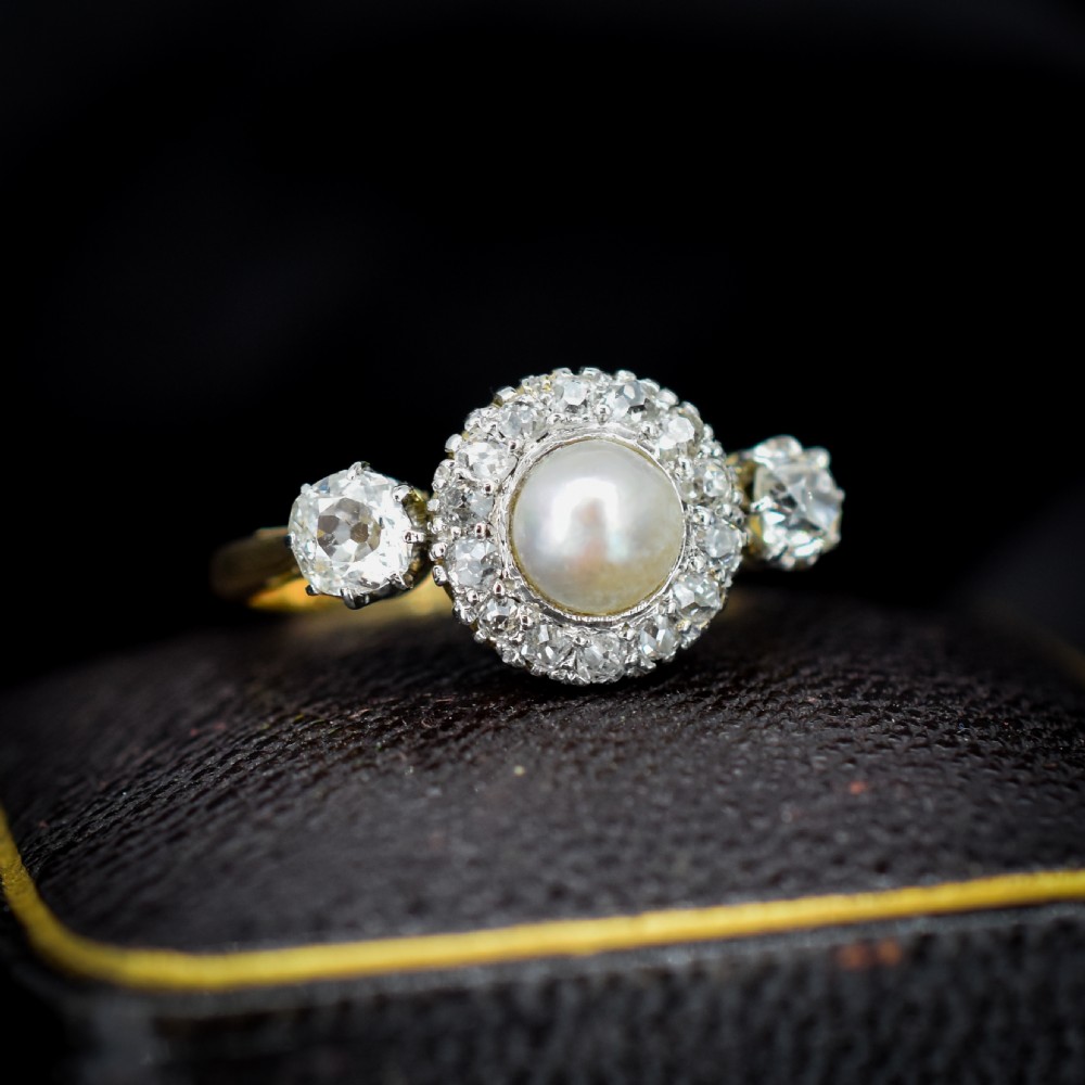 An Edwardian 18ct yellow gold ring featuring a central round pearl framed by a halo of small diamonds and two shoulder-set diamonds.