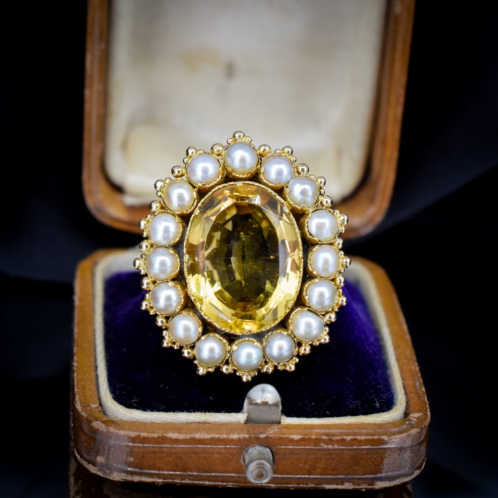 An antique yellow gold ring featuring a central oval faceted yellow citrine stone surrounded by a halo of white seed pearls.