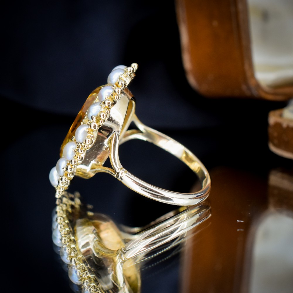 An antique yellow gold ring featuring a central oval faceted yellow citrine stone surrounded by a halo of white seed pearls. View X