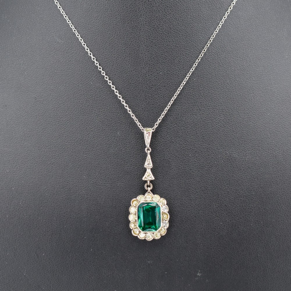 An antique silver pendant necklace featuring an emerald-cut green paste stone surrounded by a clear paste stone halo, suspended from an articulated linear drop. View II