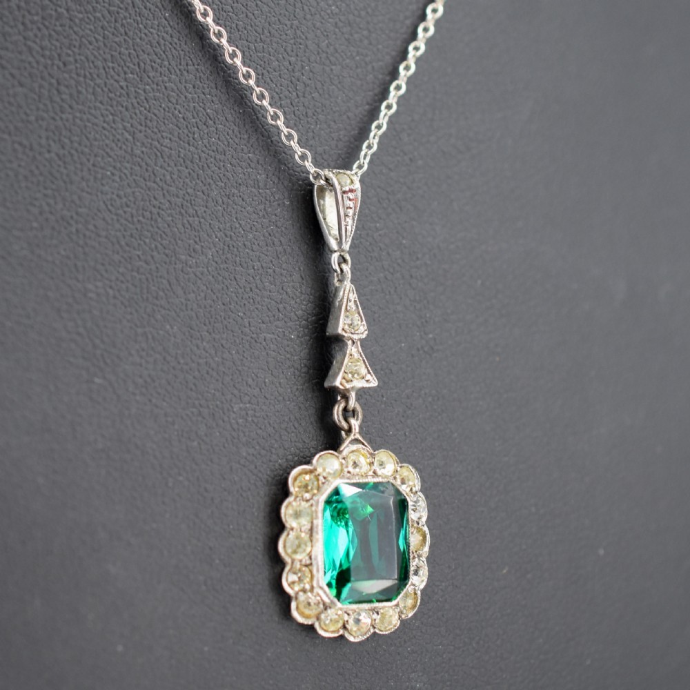 An antique silver pendant necklace featuring an emerald-cut green paste stone surrounded by a clear paste stone halo, suspended from an articulated linear drop. View III