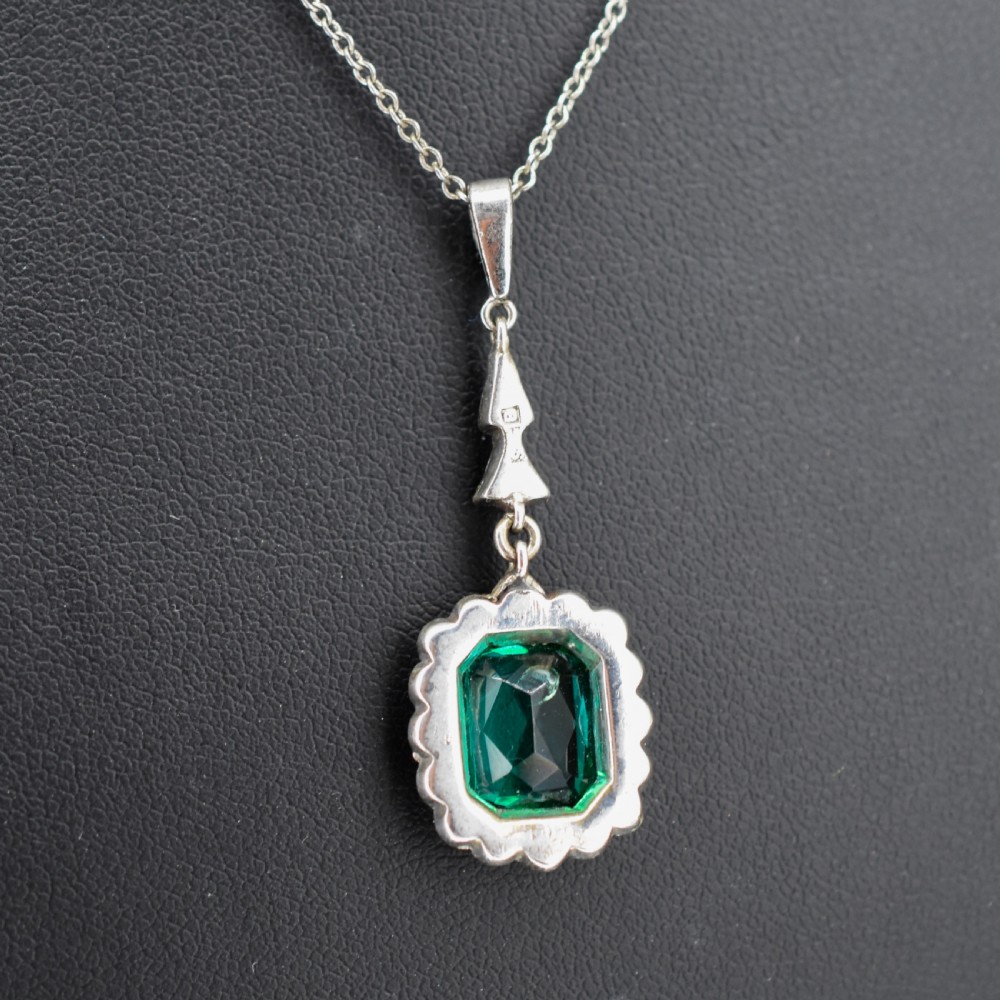 An antique silver pendant necklace featuring an emerald-cut green paste stone surrounded by a clear paste stone halo, suspended from an articulated linear drop. View V