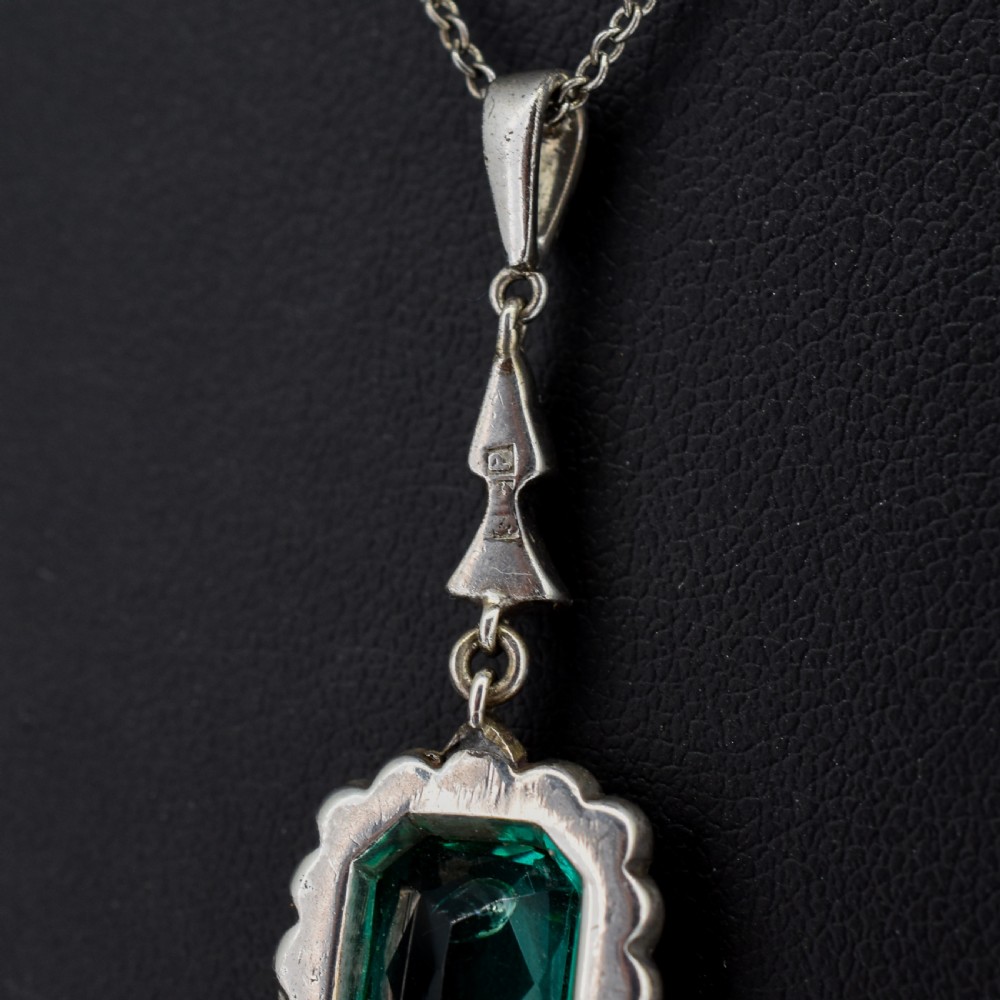 An antique silver pendant necklace featuring an emerald-cut green paste stone surrounded by a clear paste stone halo, suspended from an articulated linear drop. View VI