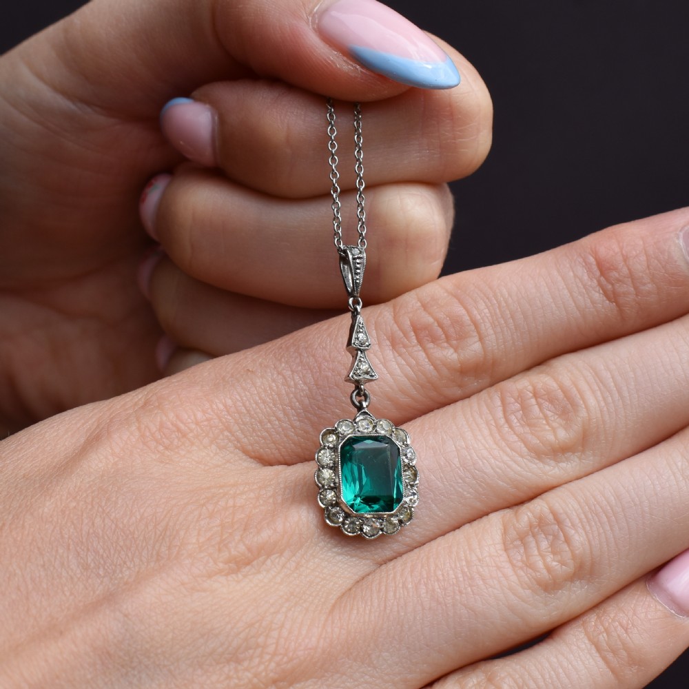 An antique silver pendant necklace featuring an emerald-cut green paste stone surrounded by a clear paste stone halo, suspended from an articulated linear drop. View VIII
