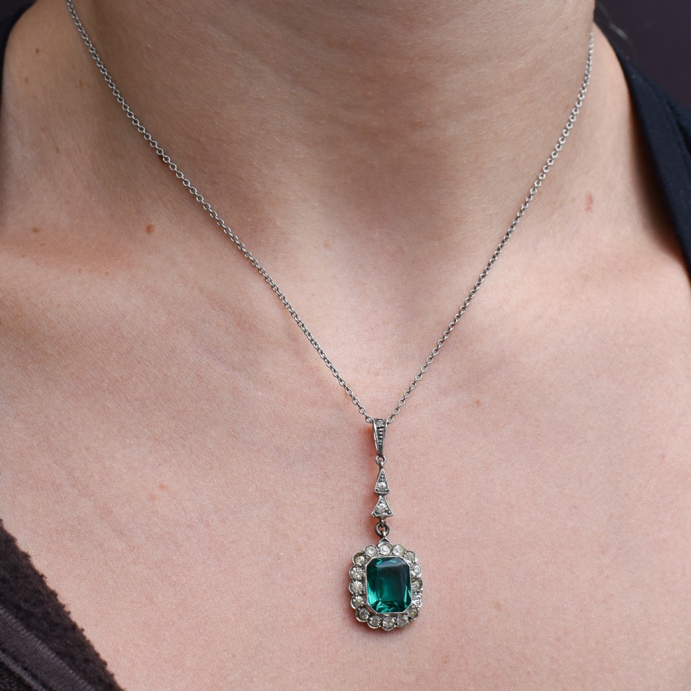 An antique silver pendant necklace featuring an emerald-cut green paste stone surrounded by a clear paste stone halo, suspended from an articulated linear drop. View IX