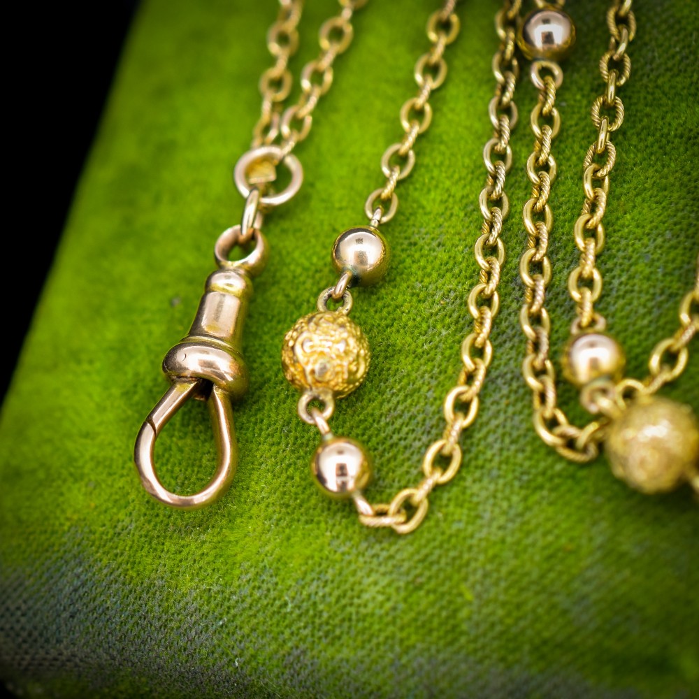 An antique Victorian 15ct yellow gold long fancy link guard chain necklace styled in multiple tiers, featuring pattern-carved gold beads and a swivel clasp hanging at the centre. View V
