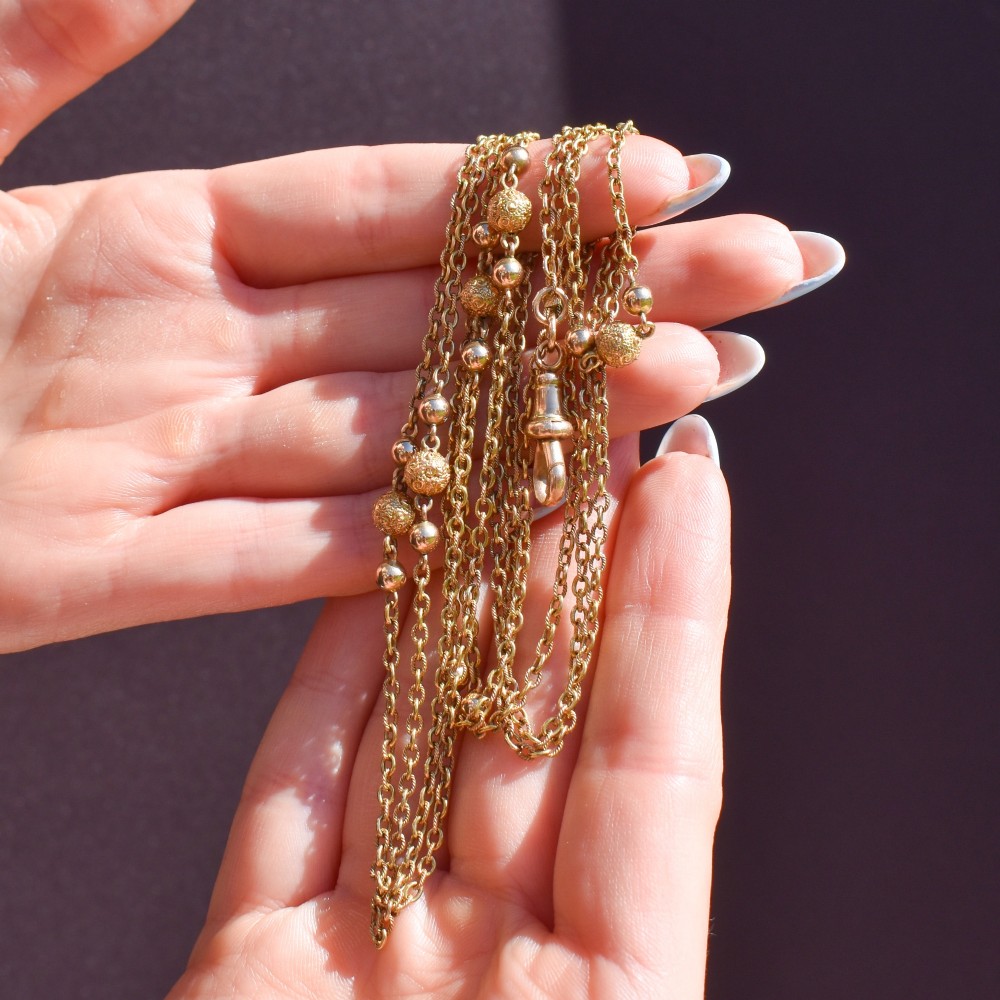 An antique Victorian 15ct yellow gold long fancy link guard chain necklace styled in multiple tiers, featuring pattern-carved gold beads and a swivel clasp hanging at the centre. View XI