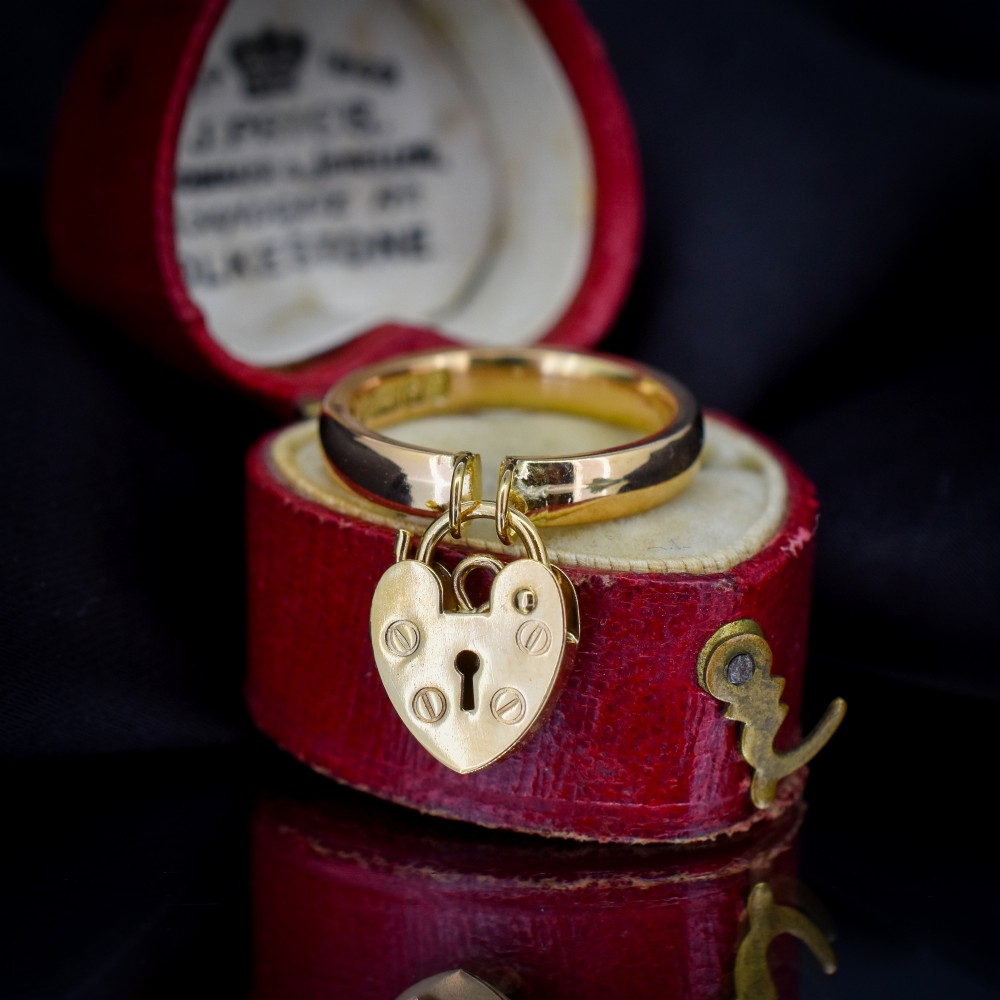 A polished 9ct yellow gold band ring featuring a suspended heart-shaped padlock drop charm with keyhole detail.
