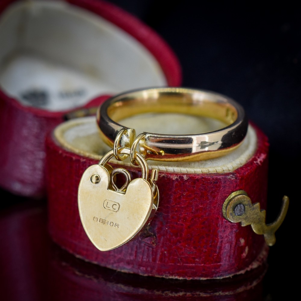 A polished 9ct yellow gold band ring featuring a suspended heart-shaped padlock drop charm with keyhole detail. View VII