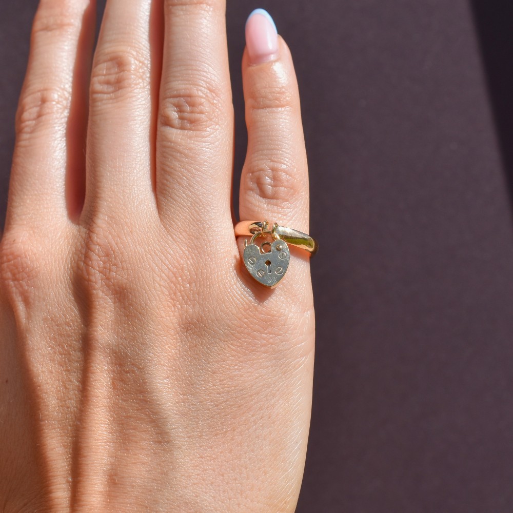 A polished 9ct yellow gold band ring featuring a suspended heart-shaped padlock drop charm with keyhole detail. View IX