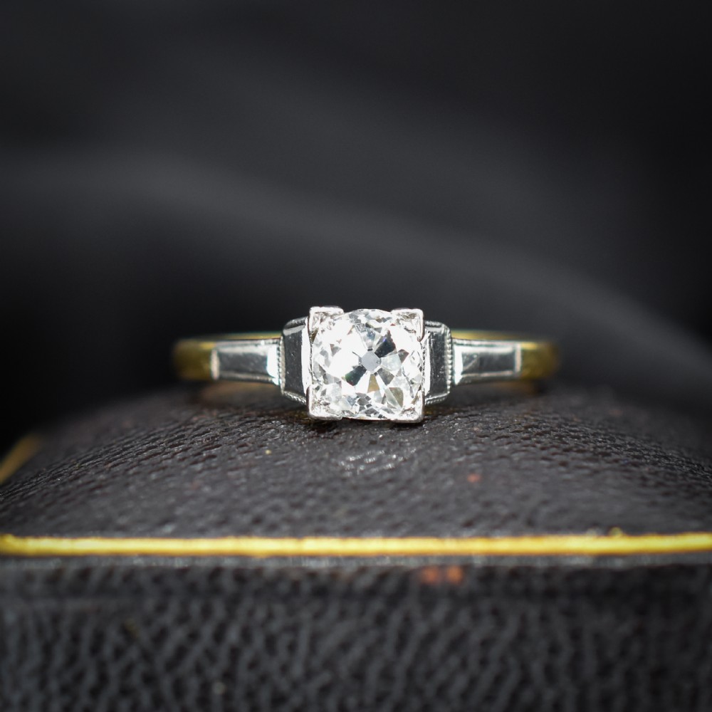 An Art Deco engagement ring featuring a square-set central old cut diamond on an 18ct yellow gold band with white metal stepped shoulders.
