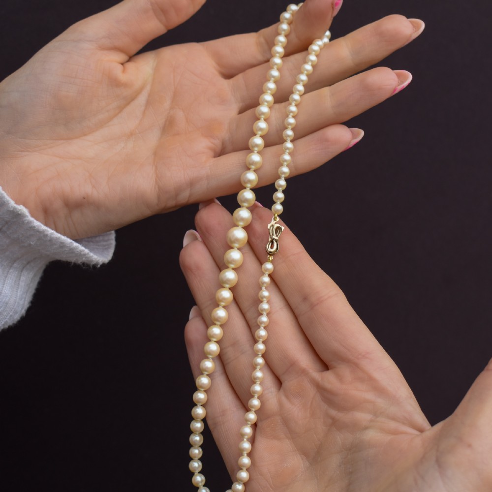 A 17-inch strand of graduated cream pearls featuring individually knotted beads and a gold bow-shaped clasp, resting on green velvet. View IX