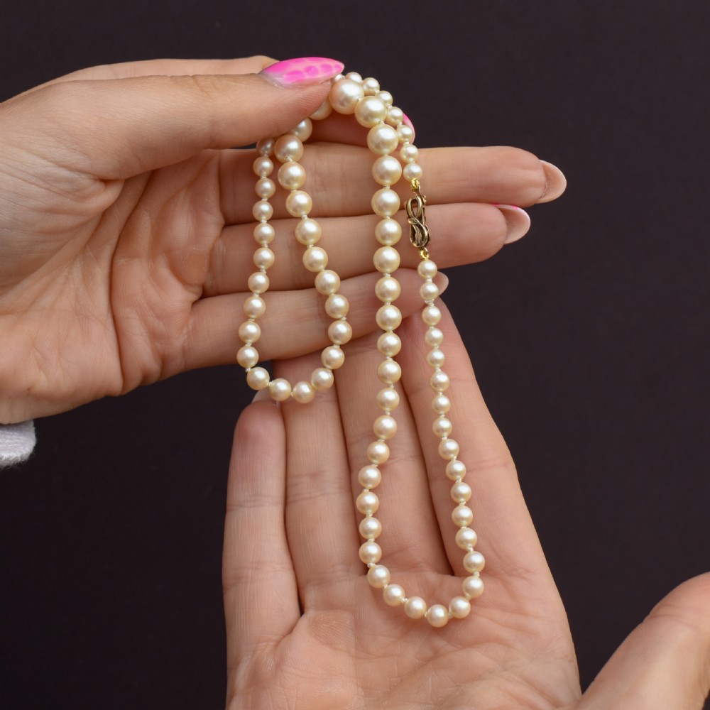 A 17-inch strand of graduated cream pearls featuring individually knotted beads and a gold bow-shaped clasp, resting on green velvet. View X