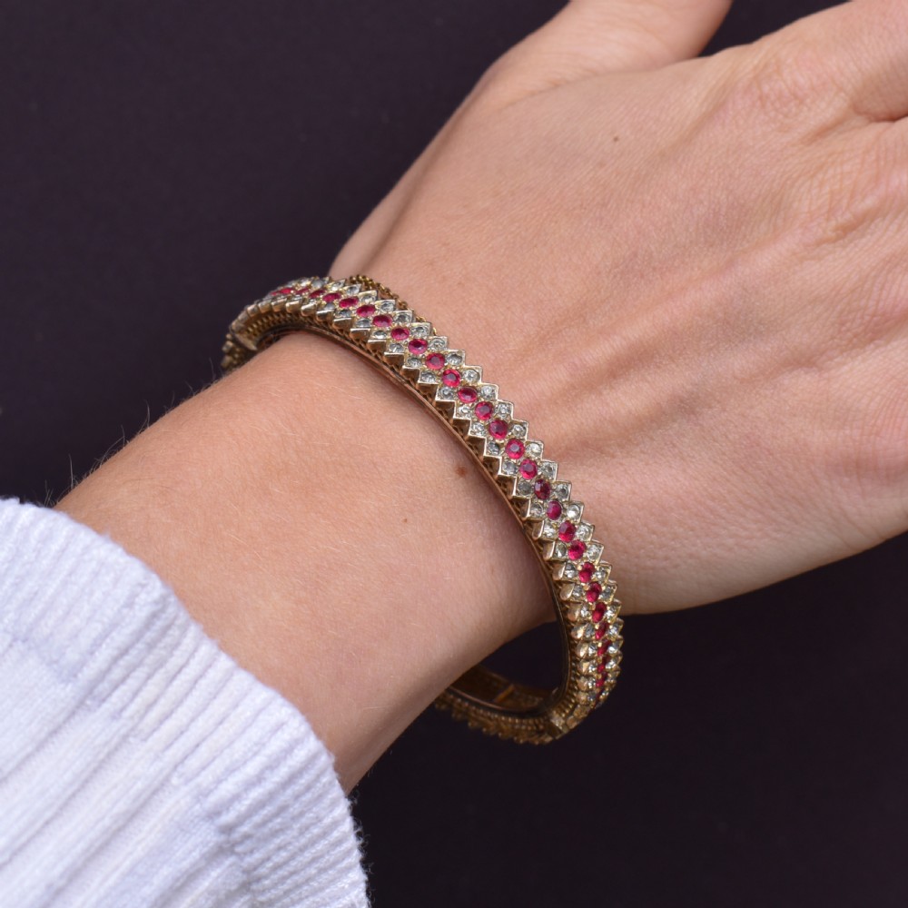 An Art Deco hinged bangle featuring a geometric chevron pattern set with alternating rows of clear and red round-cut paste stones. View X