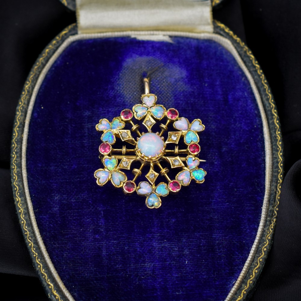 A gold starburst-shaped brooch and pendant set with a central cabochon opal surrounded by rubies, diamonds, and heart-shaped opal petals.