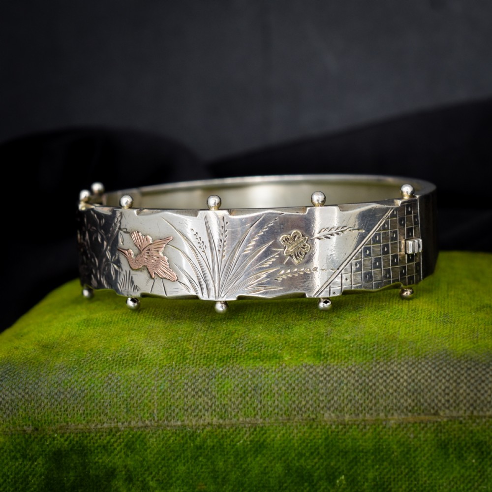 A wide silver hinged cuff bangle featuring engraved floral foliage and applied gold bird motifs along the outer band. View II