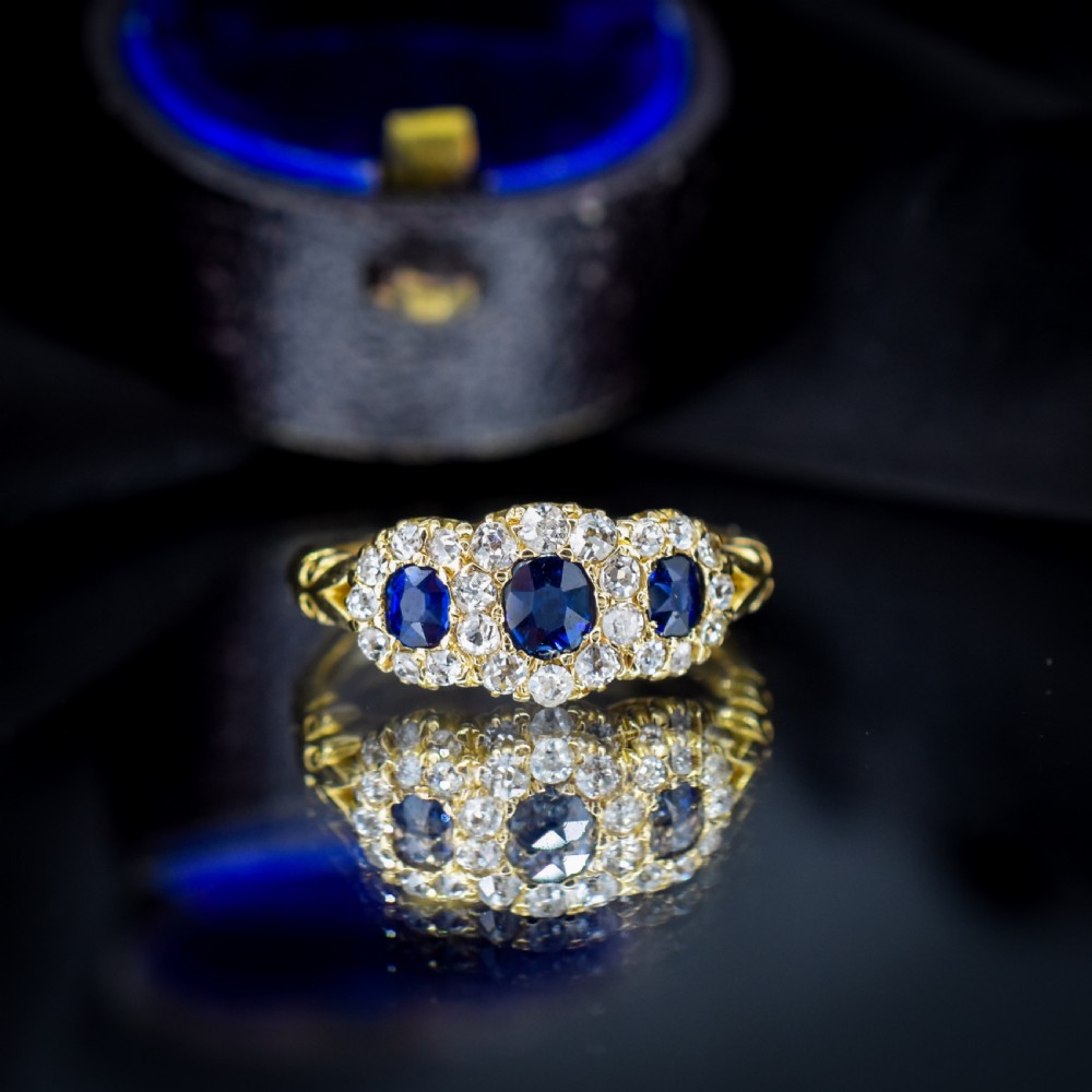 An 18ct yellow gold Victorian ring featuring three oval blue sapphires, each encircled by a cluster of small diamonds. View IV