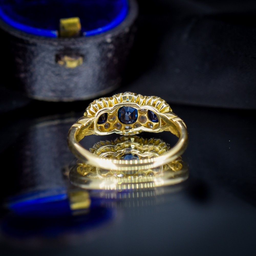 An 18ct yellow gold Victorian ring featuring three oval blue sapphires, each encircled by a cluster of small diamonds. View V