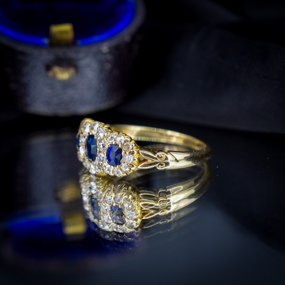 An 18ct yellow gold Victorian ring featuring three oval blue sapphires, each encircled by a cluster of small diamonds. View VI