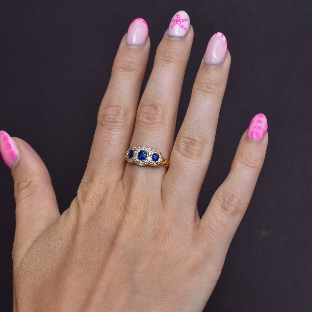 An 18ct yellow gold Victorian ring featuring three oval blue sapphires, each encircled by a cluster of small diamonds. View IX