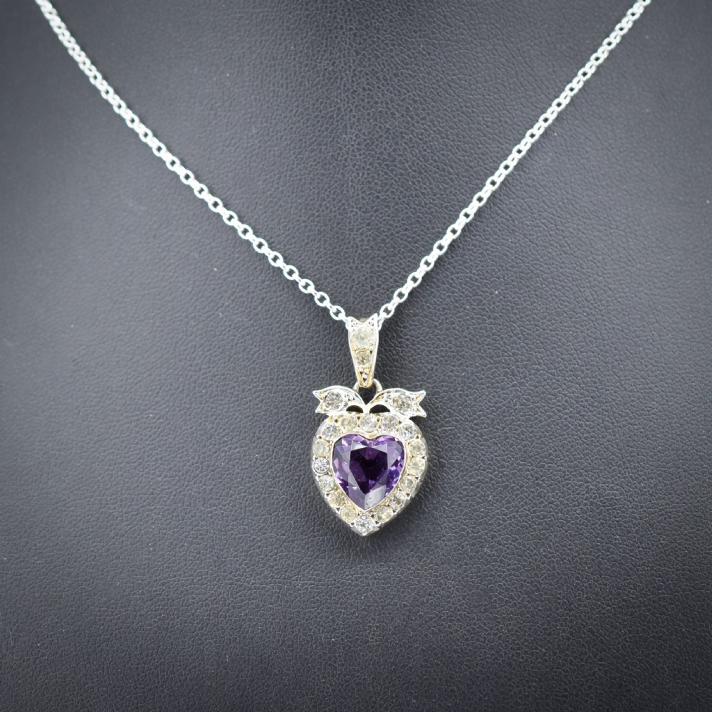 An antique silver necklace featuring a heart-shaped purple paste central stone surrounded by clear paste stones and topped with a bow motif.