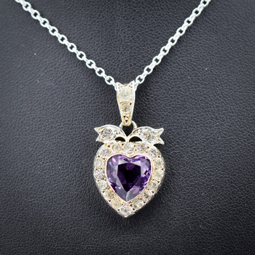 An antique silver necklace featuring a heart-shaped purple paste central stone surrounded by clear paste stones and topped with a bow motif. View II
