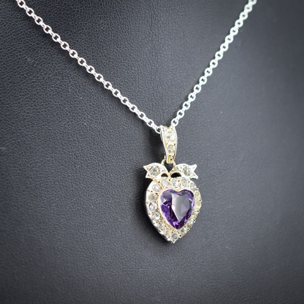 An antique silver necklace featuring a heart-shaped purple paste central stone surrounded by clear paste stones and topped with a bow motif. View III