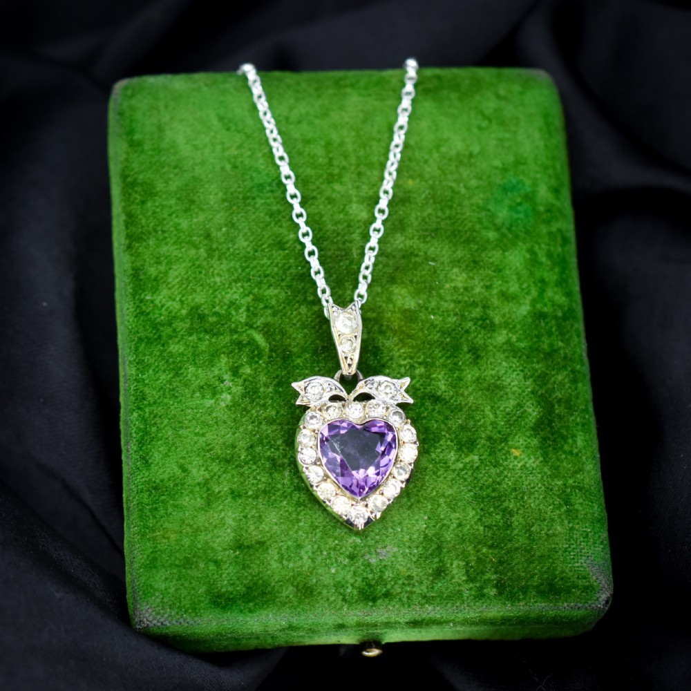 An antique silver necklace featuring a heart-shaped purple paste central stone surrounded by clear paste stones and topped with a bow motif. View VI