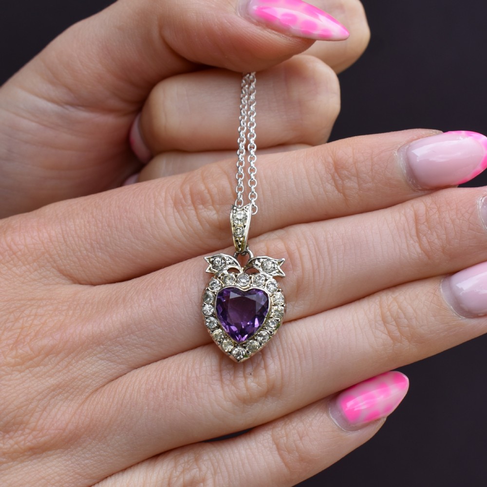 An antique silver necklace featuring a heart-shaped purple paste central stone surrounded by clear paste stones and topped with a bow motif. View VII