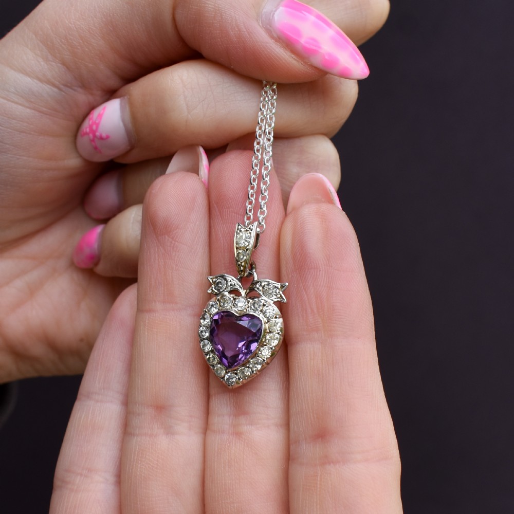 An antique silver necklace featuring a heart-shaped purple paste central stone surrounded by clear paste stones and topped with a bow motif. View VIII