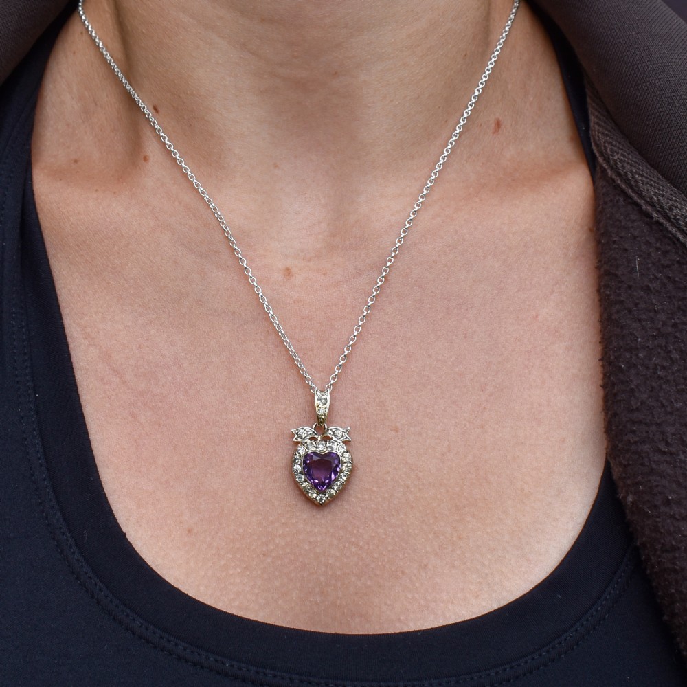 An antique silver necklace featuring a heart-shaped purple paste central stone surrounded by clear paste stones and topped with a bow motif. View IX