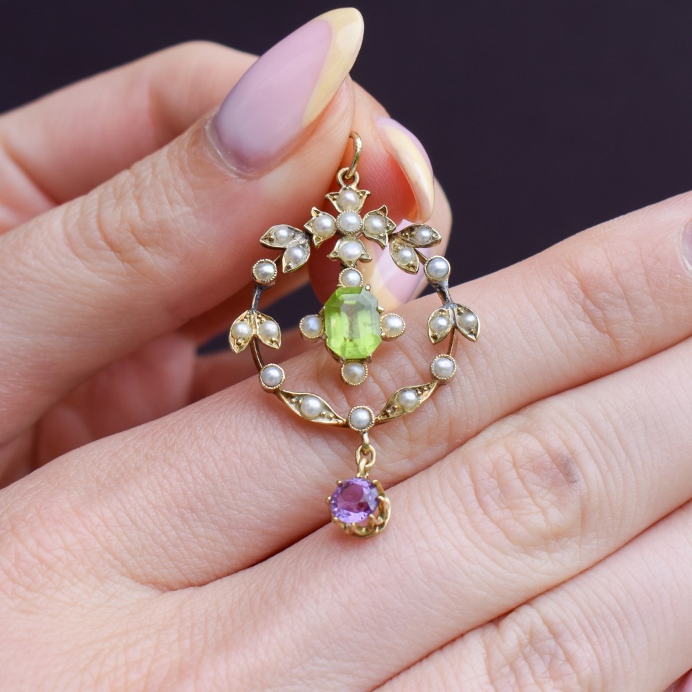 An Edwardian 15ct gold openwork lavalier pendant set with seed pearls, featuring an oval green peridot central stone and a suspended round purple amethyst drop. View VI