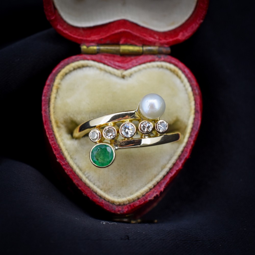 A 14ct yellow gold bypass twist ring set with a single pearl, a round green emerald, and five bezel-set diamonds across the band.