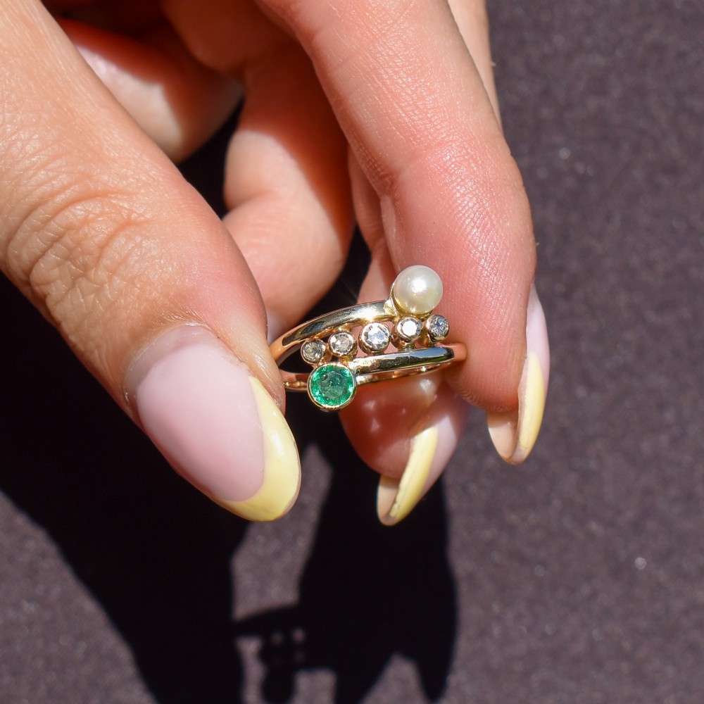 A 14ct yellow gold bypass twist ring set with a single pearl, a round green emerald, and five bezel-set diamonds across the band. View V