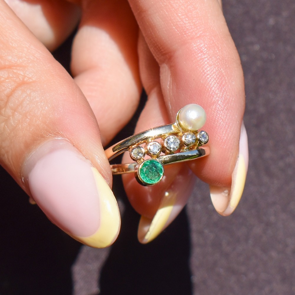 A 14ct yellow gold bypass twist ring set with a single pearl, a round green emerald, and five bezel-set diamonds across the band. View VI