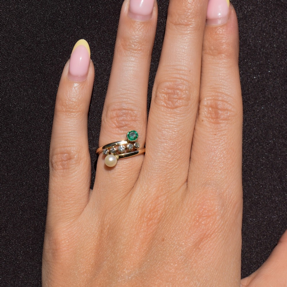 A 14ct yellow gold bypass twist ring set with a single pearl, a round green emerald, and five bezel-set diamonds across the band. View VII
