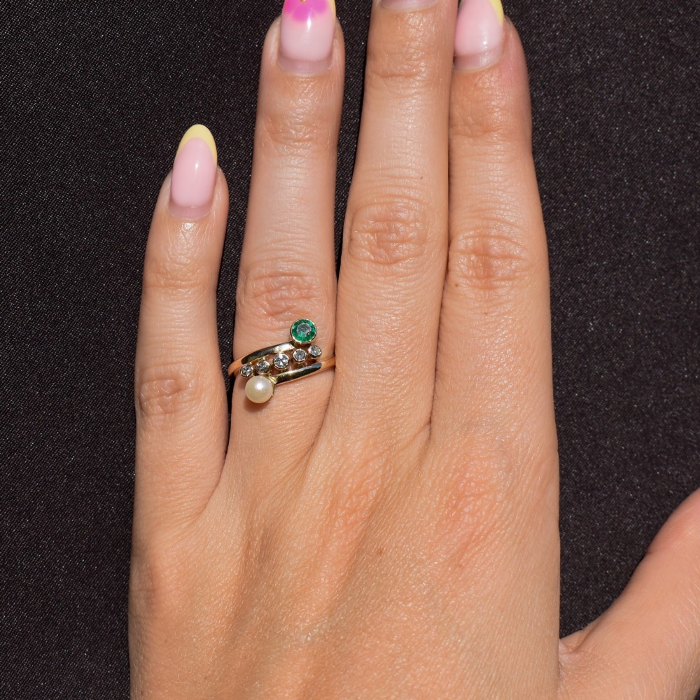 A 14ct yellow gold bypass twist ring set with a single pearl, a round green emerald, and five bezel-set diamonds across the band. View VIII