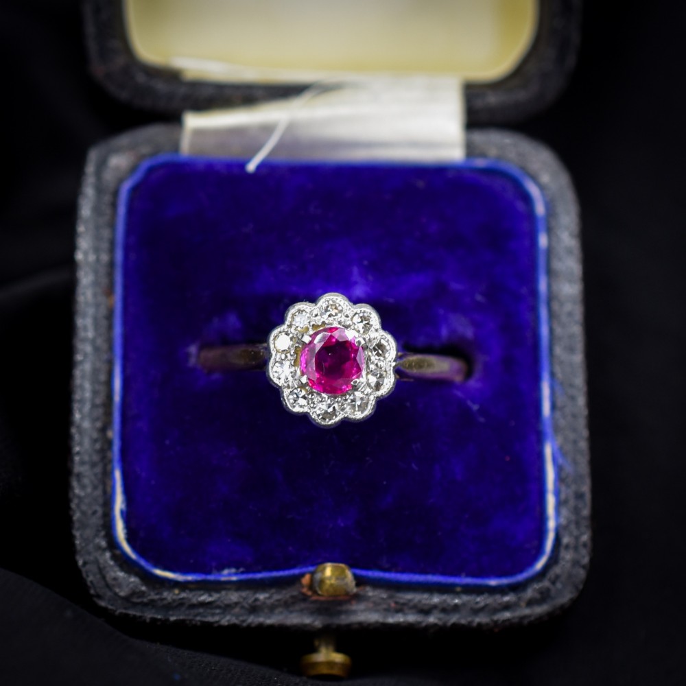 An Edwardian 18ct gold ring featuring a central oval faceted red ruby surrounded by a halo of small diamonds in a scalloped cluster setting.