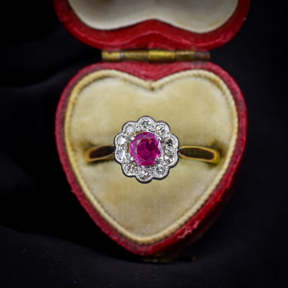 An Edwardian 18ct gold ring featuring a central oval faceted red ruby surrounded by a halo of small diamonds in a scalloped cluster setting. View II