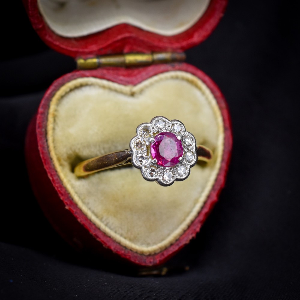 An Edwardian 18ct gold ring featuring a central oval faceted red ruby surrounded by a halo of small diamonds in a scalloped cluster setting. View III